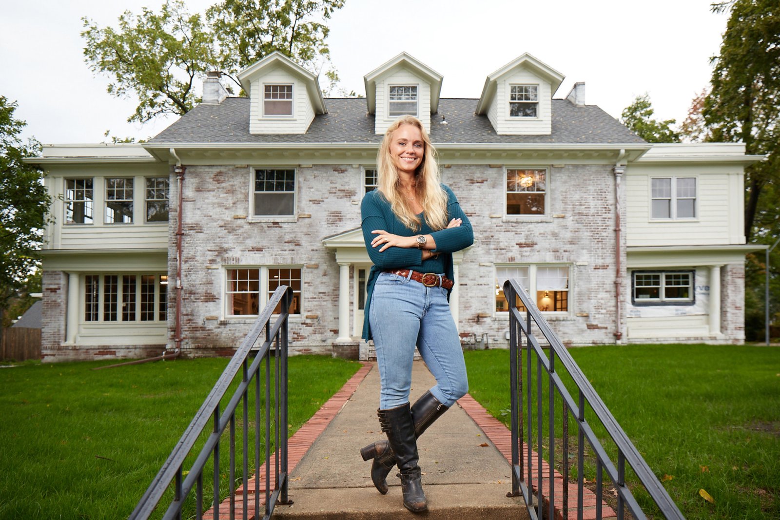 Blonde lady wearing blue jeans and a blue top stands in front of a large grey American mansion